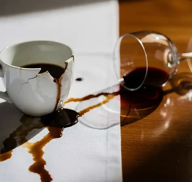 Gebarsten koffiekopje lekt espresso op wit linnen tafelkleed, met omgevallen wijnglas op gepolijst houten restauranttafel.