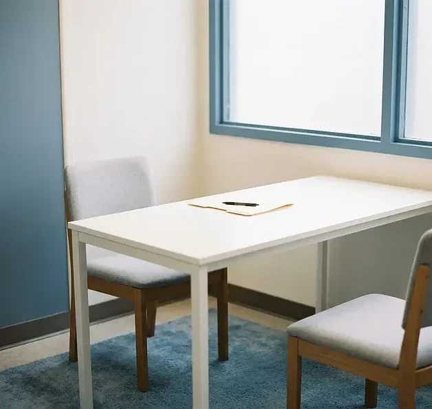 Lege mediationruimte met twee stoelen aan een witte tafel, gesloten map en pen, zacht natuurlijk licht door matglas.
